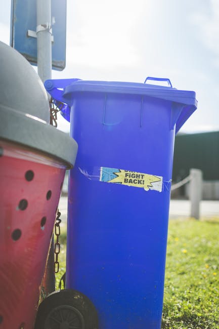 A row of large, wheeled rubbish bins aligned along a pavement next to a brick wall, with the front green bin slightly opened, showing a textured, ribbed lid with a matte finish. Behind it, blue bins w